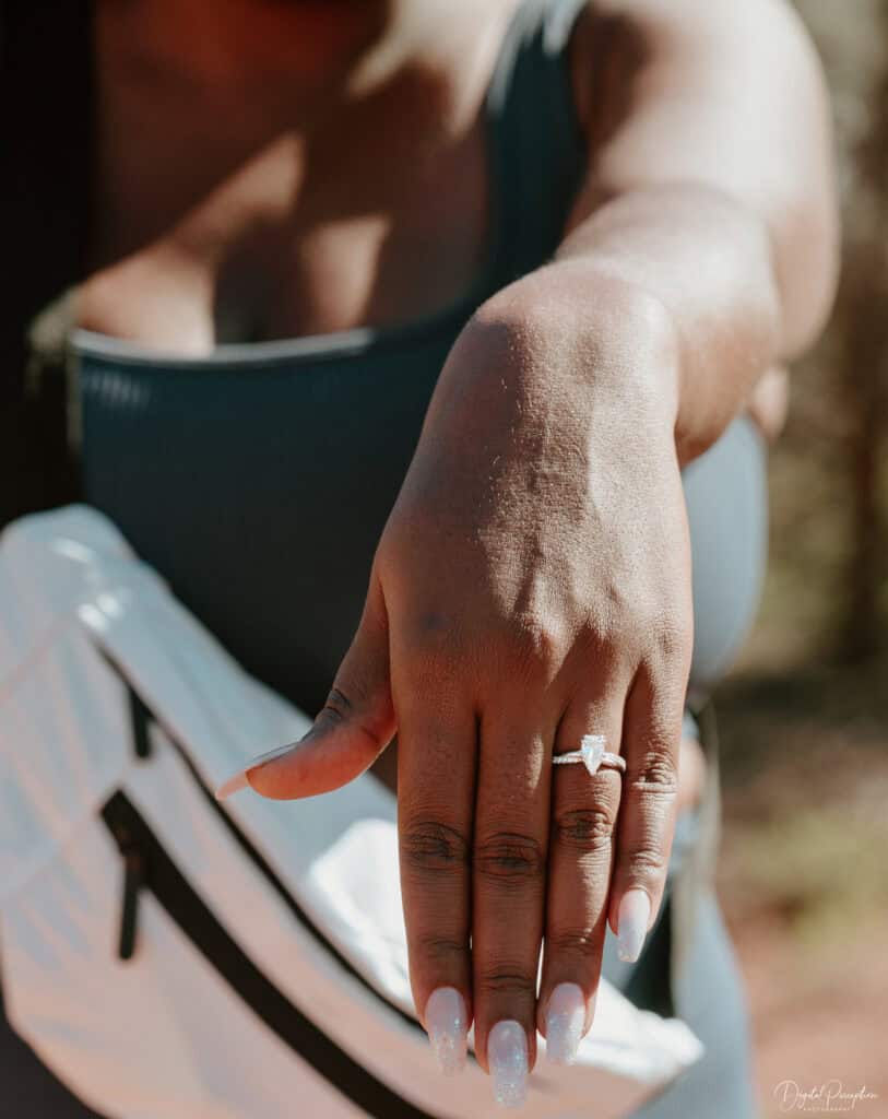 A close-up of a woman showcasing her engagement ring on her outstretched hand. She wears a blue sleeveless top and has long, glittery manicured nails. A white bag with black accents is slung over her shoulder, and the background is blurred. - Sedona, AZ