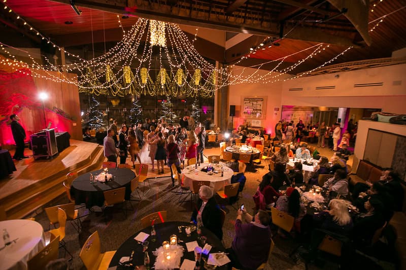 A wedding reception with guests dancing under string lights and chandeliers inside a venue with a stage and round tables.