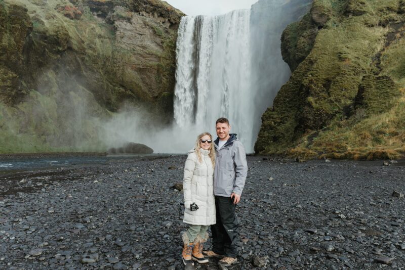 A couple stands in front of a majestic waterfall, with the man wearing a light gray jacket and dark pants, and the woman dressed in a long white coat and sunglasses, both smiling. The waterfall cascades powerfully down a rugged cliff, creating a misty spray around them, set against a backdrop of green moss-covered slopes.